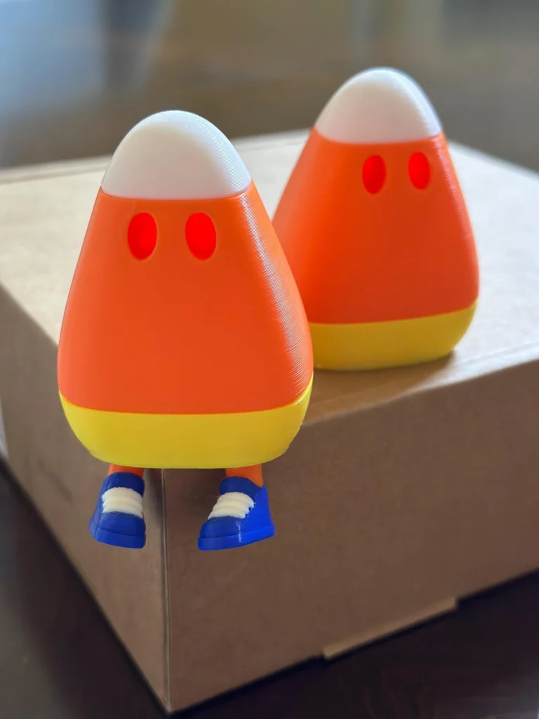 Two candy corn figures with hidden legs, in orange socks and blue shoes, sitting on the edge of a box