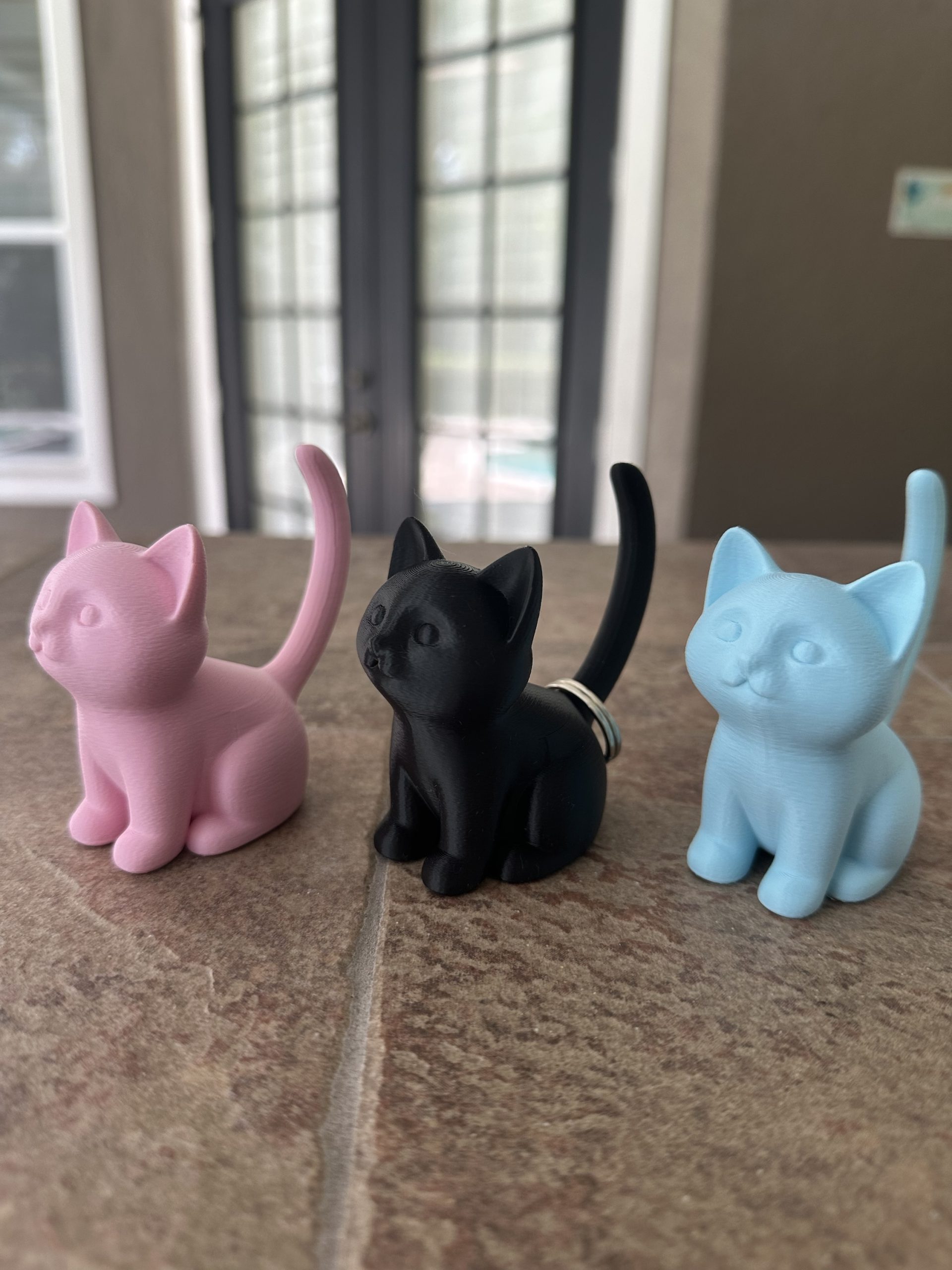 Cat ring holders in light pink, black and light blue, with a ring on the black cat’s tail